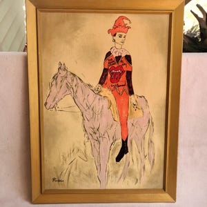 May include: Framed artwork featuring a person in a jester costume riding a horse. The person's costume is orange and black with a Rolling Stones logo. The artwork is in a gold frame.