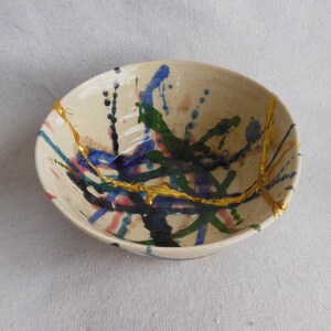 May include: A white ceramic bowl with a colorful abstract design. The bowl has splashes of blue, green, pink, and black paint, and gold accents.