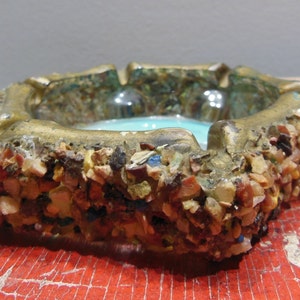 May include: A vintage ashtray made of glass and decorated with small stones. The ashtray is a light blue color and has a gold rim. The stones are a variety of colors, including brown, white, and red.