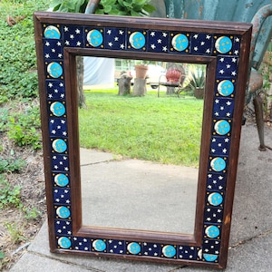 May include: Rectangular mirror with a dark brown wooden frame. The frame is decorated with blue tiles featuring a crescent moon and stars design. The mirror reflects a green lawn and outdoor setting.