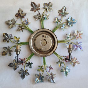 May include: A metal wall sconce with a circular backplate and a design of green, blue, and purple leaves. The sconce has two candle-style light fixtures.