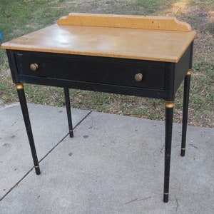Pu&ograve; includere: Un tavolo vintage nero con un solo cassetto e dettagli dorati. Il tavolo ha un piano in legno chiaro e poggia su quattro gambe.