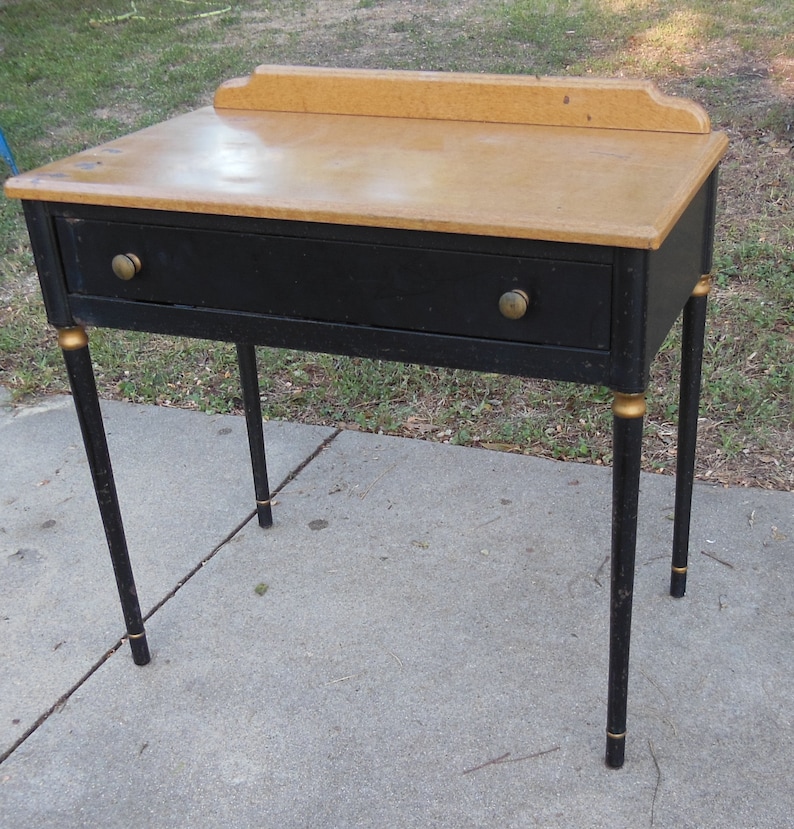 Pu&ograve; includere: Un tavolo vintage nero con un solo cassetto e dettagli dorati. Il tavolo ha un piano in legno chiaro e poggia su quattro gambe.
