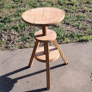 May include: A wooden adjustable stool with a round seat and three legs. The stool has a screw mechanism to adjust the height.