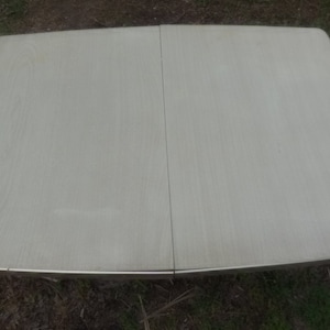 May include: A light-colored, rectangular table with a wood-grain pattern. The table has a metal trim and is approximately 36 inches wide and 48 inches long when fully extended. The table is outdoors, with a yellow chair visible.