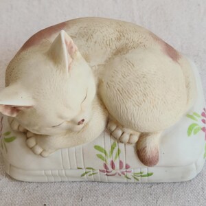 May include: A ceramic figurine of a sleeping cat. The cat is cream-colored with pink markings and is curled up on a white pillow decorated with pink and green floral designs. The cat's eyes are closed, and its paws are visible.