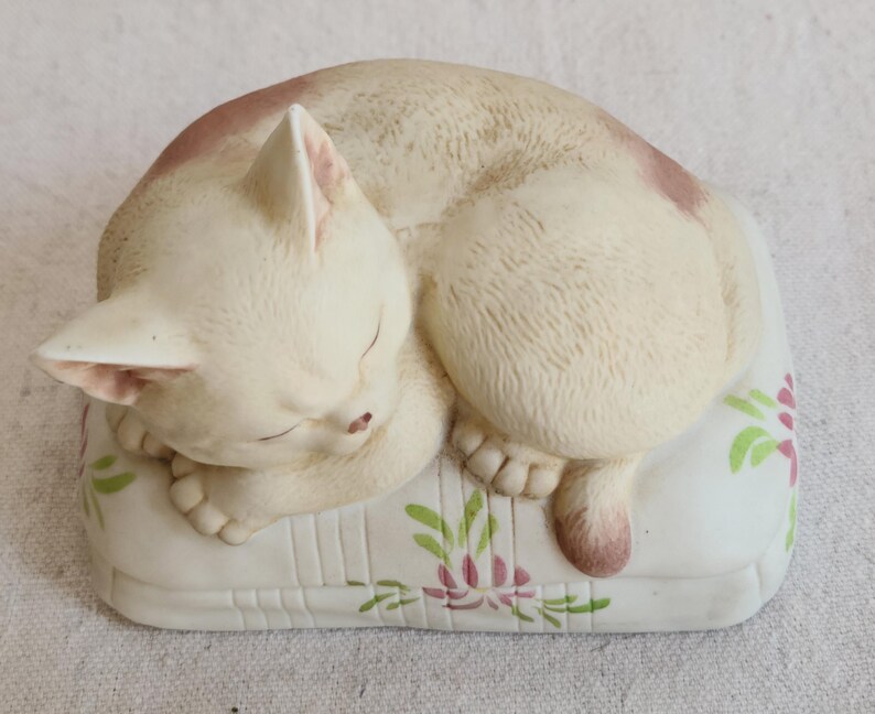 May include: A ceramic figurine of a sleeping cat. The cat is cream-colored with pink markings and is curled up on a white pillow decorated with pink and green floral designs. The cat's eyes are closed, and its paws are visible.