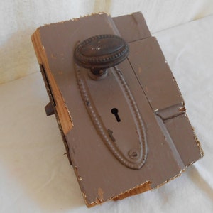 May include: A vintage door knob and lock plate with an oval-shaped keyhole and a decorative border. The knob is made of dark metal and has a rounded top. The plate is attached to a piece of wood that is weathered and has some damage.