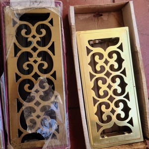 May include: Two decorative brass-colored vent covers with ornate, openwork designs. One is still in its original packaging, while the other rests in a cardboard box. The designs feature swirling patterns and geometric shapes.
