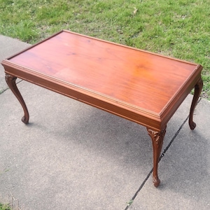 May include: A rectangular, wooden coffee table with a reddish-brown finish. The table features ornate, curved legs and a detailed edge. The table measures approximately 48 inches long and 24 inches wide.