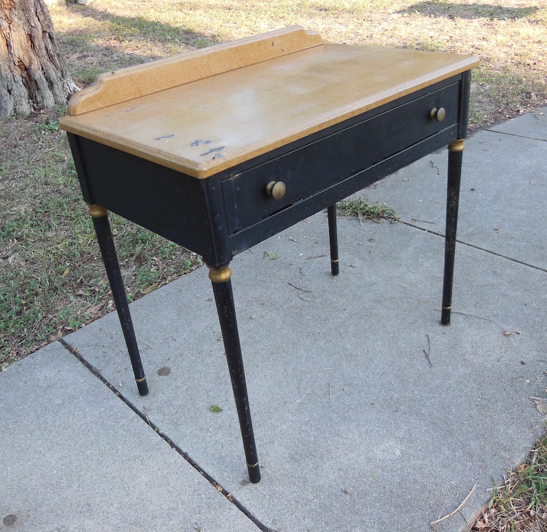 Pu&ograve; includere: Una scrivania in metallo nero con un solo cassetto e dettagli dorati. La scrivania ha un piano in legno chiaro e poggia su quattro gambe.