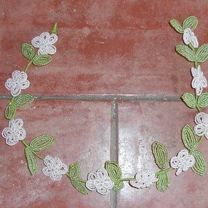 May include: A white and green beaded garland with white flowers and green leaves. The garland is shaped like a circle and is lying on a red tiled floor.