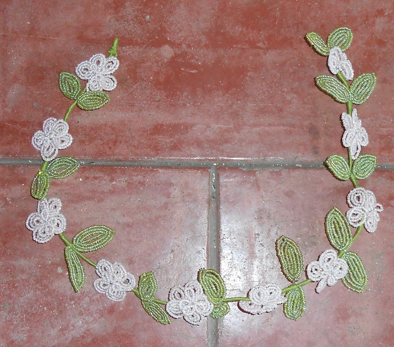 May include: A white and green beaded garland with white flowers and green leaves. The garland is shaped like a circle and is lying on a red tiled floor.