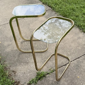 May include: Two vintage side tables. Each table has a clear glass top and a gold-colored metal frame. The frame has a unique, curved design. The tables are placed on a concrete surface.