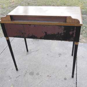 Pu&ograve; includere: Una scrivania in legno vintage con gambe verniciate di nero e dettagli dorati. La scrivania ha una finitura rosso scuro e nera con alcuni segni di usura.