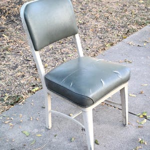 May include: A vintage chair featuring a white metal frame and a distressed, green vinyl seat and back. The chair exhibits signs of wear, with scratches and peeling on the seat. The chair is positioned on a concrete surface outdoors.
