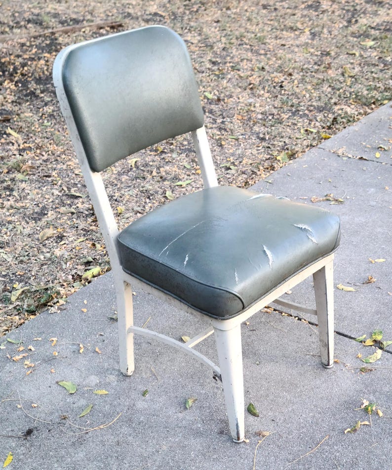 May include: A vintage chair featuring a white metal frame and a distressed, green vinyl seat and back. The chair exhibits signs of wear, with scratches and peeling on the seat. The chair is positioned on a concrete surface outdoors.