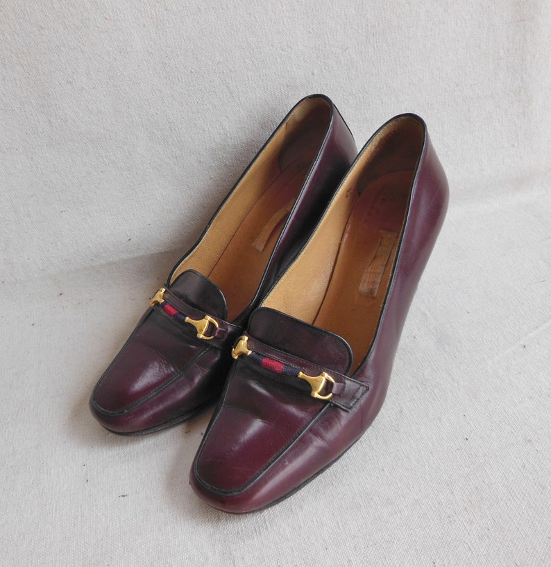 May include: A pair of burgundy leather heeled shoes with gold buckles and a red, white, and blue ribbon detail.