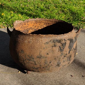 May include: A large, weathered, rusty metal cauldron with two curved handles. The cauldron has a wide, irregular rim and a rounded body. The exterior shows signs of age and use, with dark patches and a rough texture. The cauldron sits on a concrete surface.