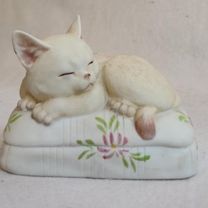 May include: A cream-colored ceramic cat figurine, sleeping peacefully on a white pillow. The pillow is decorated with pink and green floral designs. The cat's eyes are closed, and its paws are tucked in.