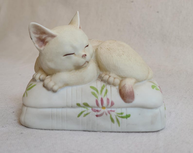 May include: A cream-colored ceramic cat figurine, sleeping peacefully on a white pillow. The pillow is decorated with pink and green floral designs. The cat's eyes are closed, and its paws are tucked in.