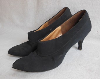 May include: A pair of black heeled shoes with a pointed toe and a fabric upper. The shoes have a simple design and a classic silhouette.