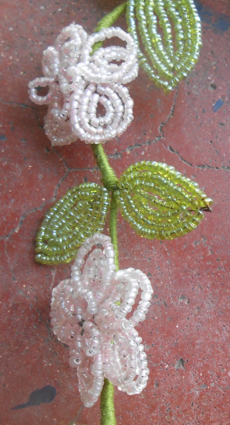 May include: A beaded flower garland with white flowers and green leaves. The garland is made of small, round beads and is strung together on a green thread.
