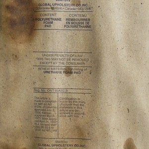 May include: A brown paper tag with white text, indicating that the product contains new material. The tag is labeled "New Material Only" and includes the company name "Global Upholstery Co. Inc." and the address "560 Supertest Road, Downsview Ontario M3J 2M6". The tag also includes the text "Under Penalty of Law This Tag May Not Be Removed Except By The Consumer" and "All New Material Consisting of Urethane Foam Pad".