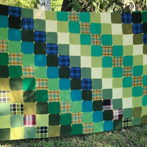 May include: A patchwork quilt with a grid pattern of green squares. The squares vary in shades of green, with some featuring plaid patterns in green, blue, and yellow. The quilt is hanging outdoors, showcasing its handmade quality.