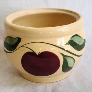May include: A cream-colored ceramic bowl with a wide opening. The bowl is decorated with a large burgundy-colored apple and green leaves. The design is painted on the exterior of the bowl.