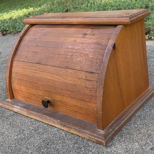 May include: A medium-toned wooden bread box featuring a curved roll-top door with a small black circular knob. The structure has a rectangular base, flat top, and side panels with a visible wood grain texture throughout the entire exterior surface.