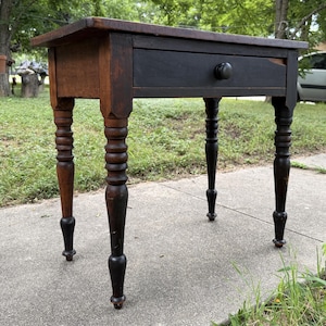 May include: A wooden side table featuring a rectangular top with a dark brown finish and a single drawer painted in black. The table stands on four turned legs that transition from a natural wood tone at the top to a dark black finish at the base.