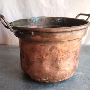 May include: A vintage copper pot with two sturdy handles. The pot has a rounded body and a wide rim, showing signs of age and use. The copper has a warm, reddish-brown hue with areas of green oxidation, adding to its antique charm.