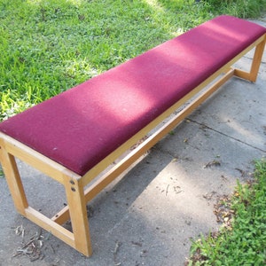 May include: A wooden bench with a burgundy fabric seat. The bench has a simple design with a rectangular frame and a flat seat.