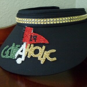 May include: Black visor with a gold beaded trim and a beaded design that says "Golf Aholic" with a red, white, and green flag.