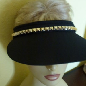 May include: A black visor with a gold studded trim. The visor is worn on a mannequin head.