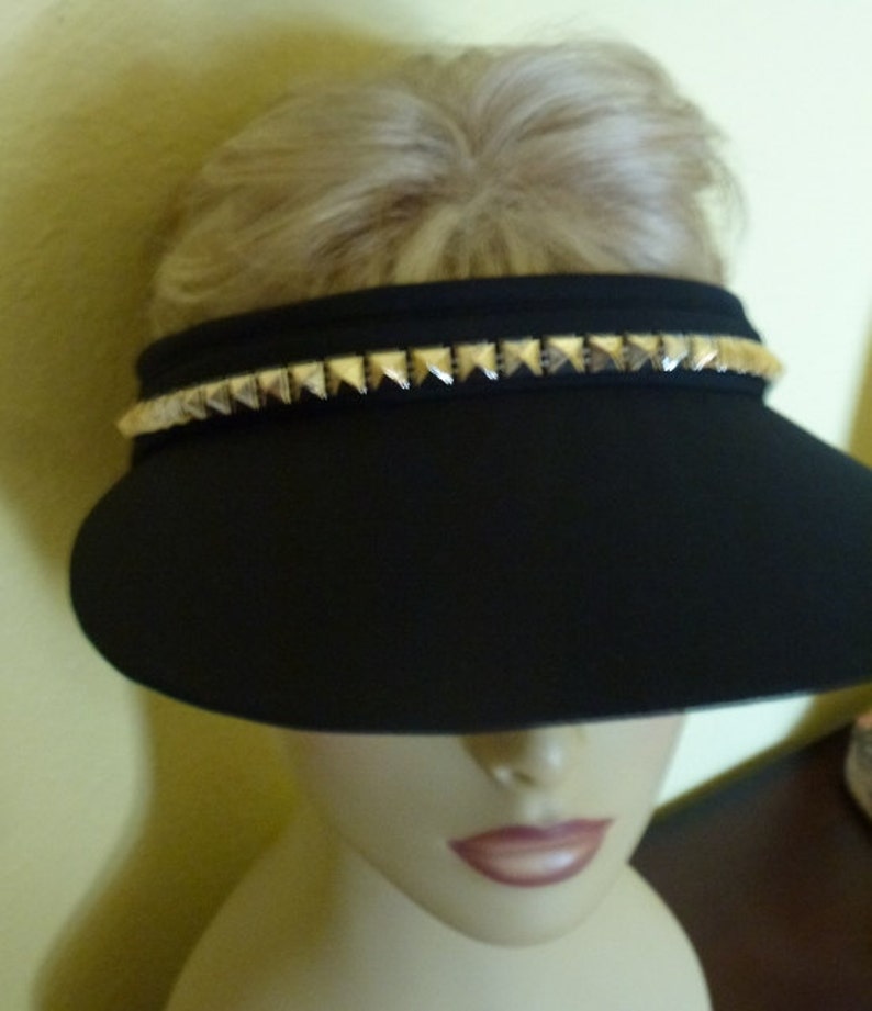 May include: A black visor with a gold studded trim. The visor is worn on a mannequin head.