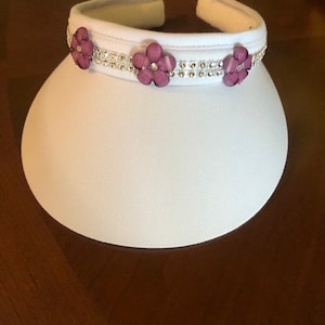 May include: White visor with a band of rhinestones and purple flower embellishments.