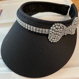 May include: A black visor with a silver rhinestone trim and a silver rhinestone bow.