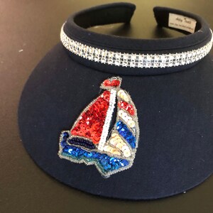 May include: A navy blue visor with a white rhinestone trim and a sailboat design made of colorful sequins.