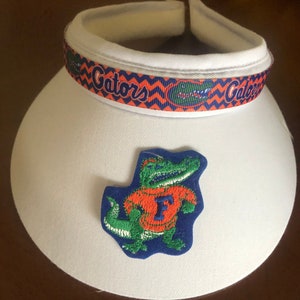 May include: White visor with a blue and orange ribbon that says "Gators" and a green alligator wearing an orange shirt with a blue "F" on it.