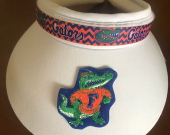 May include: White visor with a blue and orange ribbon that says "Gators" and a green alligator wearing an orange shirt with a blue "F" on it.