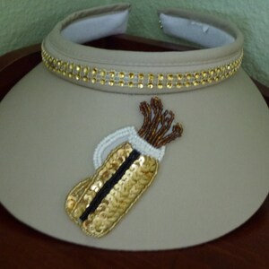 May include: A beige visor with a gold beaded trim and a gold and brown embroidered golf bag design.