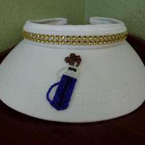 May include: White visor with a gold rhinestone trim and a beaded golf bag design.