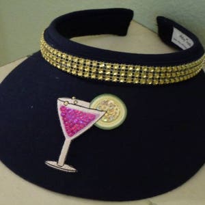 May include: A black visor with a gold rhinestone trim and a pink and white cocktail applique. The applique features a martini glass with a lime wedge.
