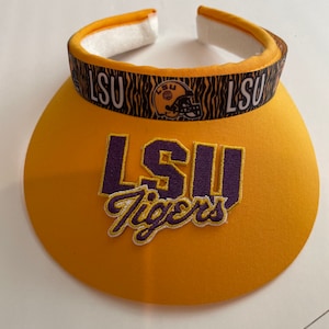 May include: A yellow visor with the text "LSU Tigers" embroidered in purple and white. The visor's band features the letters "LSU" and a tiger print design with a football helmet graphic. The visor has a white padded interior.