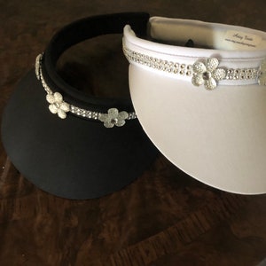 May include: Two visor hats with rhinestone trim and flower embellishments. One hat is black and the other is white.