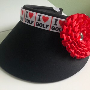 May include: Black visor with a white band that says "I love golf" with red hearts. The visor has a red fabric flower on the side.