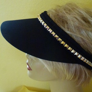 May include: A black visor with a gold studded trim. The visor is worn by a mannequin with blonde hair.