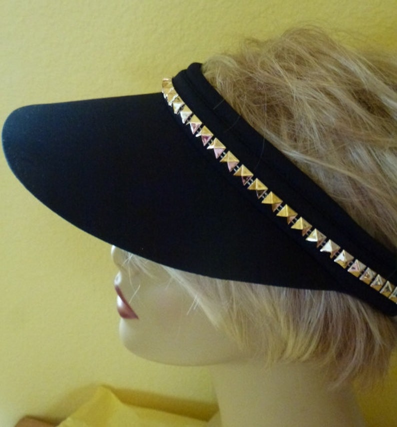 May include: A black visor with a gold studded trim. The visor is worn by a mannequin with blonde hair.